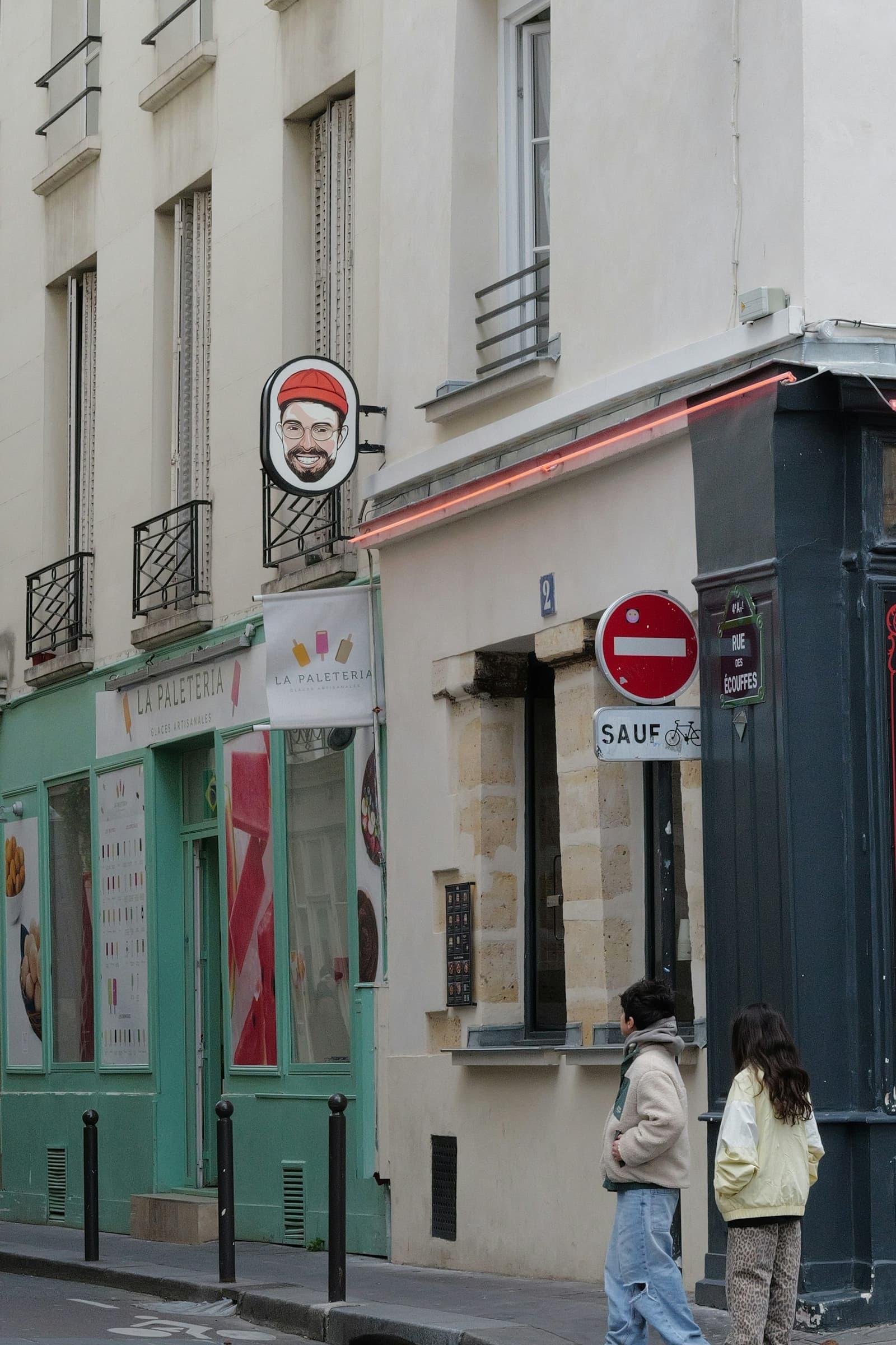 Deux passants à un coin de rue parisien, devant des commerces.